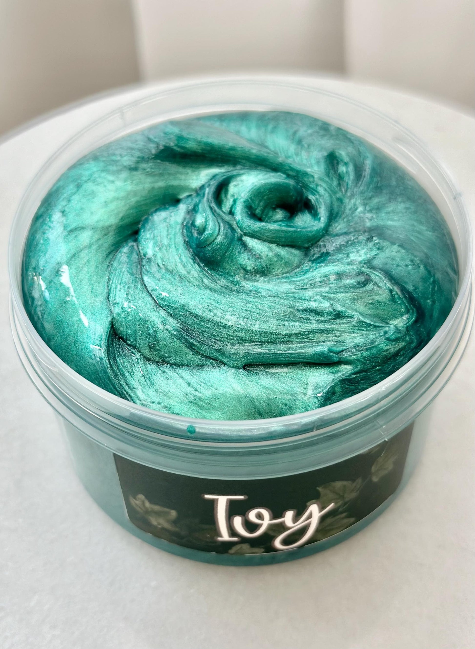 EinDunkelgrünMetallicschimmernderSlime, in einem transparentem Container. Das Etikett ist Dunkelgrün, mit Efeuranken. Es stehtdas Wort Ivy darauf.